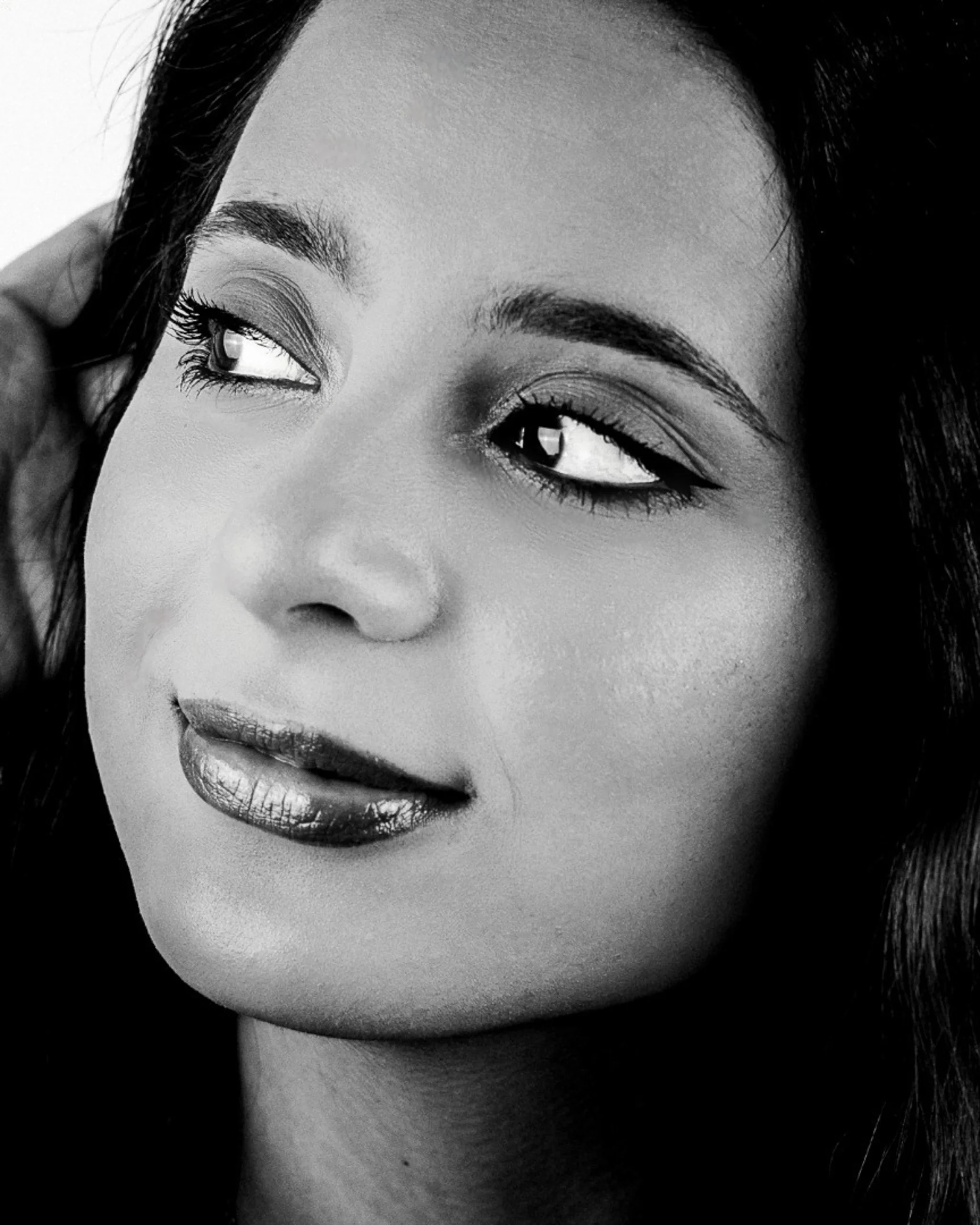 A high-contrast, black-and-white close-up portrait of a woman looking off to the side with a soft smile. The photo emphasizes her dark eyeliner and long lashes, with a clean, bright background that highlights the details of her facial features.