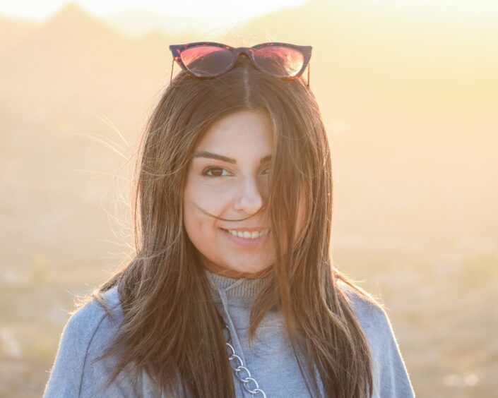 A golden hour portrait of a smiling woman with long brown hair, wearing sunglasses on her head and a grey sweater. The background is softly blurred with warm, bright sunlight creating a natural lens flare and a glowing rim light around her hair.